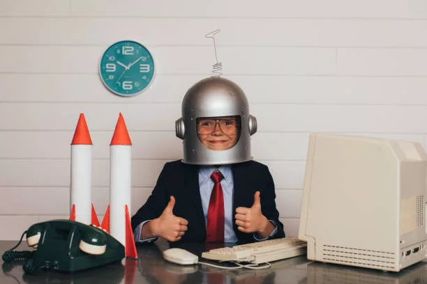 A young business boy dressed in business suit and tie sits at his office desk wearing an astronaut helmet with his two thumbs up. There are rockets on his desk as he is ready to launch his business into space and greater profitability.