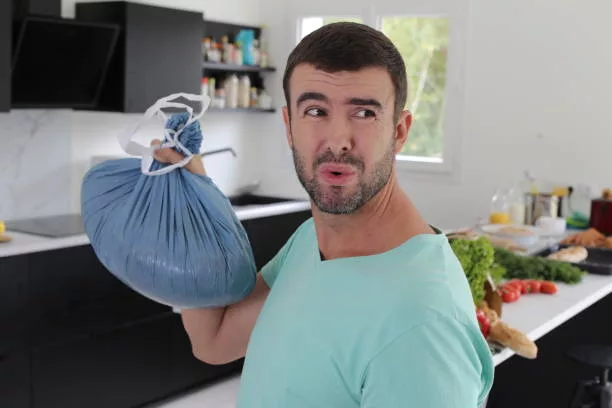 A very disgusted looking man is holding a stinky garbage bag. Looks like the residuals are rotten and creating a bad odor.